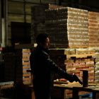 Ein Mann in Lederjacke hält eine Pistole in einem Lager mit gestapelten Kartons vor sich.