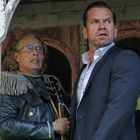 Zwei Männer schauen aufmerksam in die Ferne. Einer trägt eine beeindruckende Jacke, der andere einen Anzug. Sie stehen draußen.