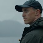 Ein Mann im Profil trägt eine dunkle Kappe und Jacke, schaut ernst zur Seite. Der Hintergrund ist neblig.