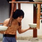 Kein Text vorhanden. Ein muskulöser Mann balanciert zwei Schüsseln und einen Stuhl auf Kopf und Schultern.