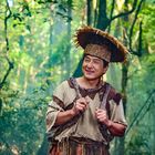 Kein Text vorhanden. Ein Mann in traditioneller Kleidung steht lächelnd in einem dichten, grünen Wald.
