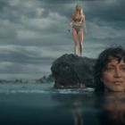 Zwei Frauen am Meer: eine schwimmt, die andere steht auf einem Felsen, bewölkter Himmel im Hintergrund.