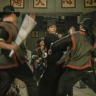 Die oberen Schilder zeigen chinesische Schriftzeichen. Unten kämpfen Männer in schwarzen Kostümen mit Hüten.