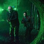 Zwei Personen mit Waffen stehen in einem grün beleuchteten Tunnel, der durch ein Gitter unterbrochen ist.