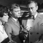 Es sind drei Personen abgebildet: zwei Männer und eine Frau, die ernsthaft auf ein Dokument blicken.