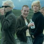 Vier junge Männer im Freien, lässig gekleidet, einer trinkt aus einer Dose, ein anderer hält eine Flasche.