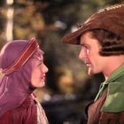 Eine Frau in einem violetten Gewand steht einem Mann mit grünem Umhang und braunem Hut mit Feder gegenüber.