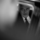 Ein älterer Mann mit Schnurrbart schaut ernst aus einem Autofenster. Es herrscht eine schwarz-weiße, melancholische Stimmung.
