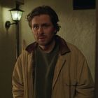 Ein Mann mit brauner Jacke und grünem Pullover steht in einem beleuchteten Flur mit altmodischer Wandlampe.