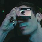 Ein junger Mann hält sich eine Kamera vors Gesicht, er steht im Dunkeln vor einem verschwommenen Hintergrund.