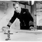 Ein Mann in einer Marineuniform beugt sich über einen Tisch mit kleinen Flaggen. Hintergrund: Karte der "GREENLAND AND BARENTS SEAS".