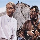 Zwei Männer in historischen Kleidern vor einem felsigen Hügel; einer hält ein Gewehr, der andere trägt eine weiße Tunika.