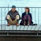 Zwei Personen sitzen an einem Steg vor einem Geländer mit Blick auf ruhiges Wasser, beide entspannt in Winterkleidung.