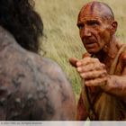 Ein Mann, stark verschmutzt, mit ausgestrecktem Arm; neben ihm eine dunkelhaarige, ebenfalls schmutzige Person in einer Wiese.