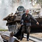 Ein älterer Mann mit weißem Haar und Bart steht vor einem alten Panzer im Wald. Er trägt ein dunkles Gewand und hohe Stiefel.