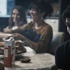 Ein Mann sitzt in einem abgedunkelten Raum, umgeben von lächelnden Menschen. Ein Getränkekarton steht auf dem Tisch.