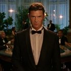 Ein Mann im Smoking steht ernst vor einem belebten Casino-Tisch. Menschen in Abendgarderobe spielen Karten.