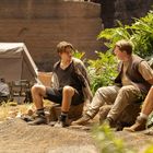 Drei Männer sitzen auf Steinen, umgeben von Pflanzen. Im Hintergrund ein Zelt mit mehreren Menschen, die beschäftigt wirken.