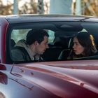 Zwei Menschen sitzen in einem roten Auto und schauen sich intensiv an. Die Frau hält das Lenkrad.