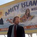 Plakat mit "Amity Island", eine Frau ruft "Help! Shark". Drei Männer stehen davor und diskutieren.