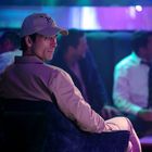 Ein Mann mit heller Jacke und Kappe sitzt in einer Bar, umgeben von anderen Personen in Anzügen, in stimmungsvollem Licht.