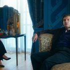 Zwei Personen in einem eleganten Raum mit blauen Wänden, einem Spiegel, Vorhängen und einer Stehlampe.