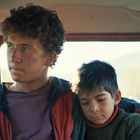 Zwei Jungen sitzen nebeneinander in einem Fahrzeug. Der ältere Junge schaut nachdenklich, während der jüngere seinen Kopf anlehnt.