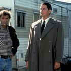 Zwei Männer stehen vor einem Wohnwagen. Einer trägt einen karierten Hemd und Mantel, der andere einen Trenchcoat und Krawatte.