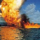 Ein riesiger Feuerball und schwarze Rauchwolken steigen aus dem Meer auf, umgeben von blauem Himmel und Wasser.