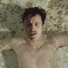 Ein Mann mit dunklem Haar liegt mit ausgestreckten Armen entspannt im flachen Wasser. Die Wasseroberfläche glitzert.