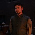 Ein nachdenklicher Mann in historischer Kleidung steht im Schein von Fackellicht vor einer dunklen Mauer.