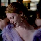 Eine Gruppe von drei Personen lehnt sich eng aneinander. Die mittlere Person trägt ein fliederfarbenes Kleid.