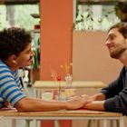 Ein Schild mit der Aufschrift "CERRADO". Zwei Männer sitzen an einem Tisch, halten sich lächelnd die Hände.