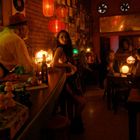 Eine gemütliche Bar mit schummriger Beleuchtung, roten Laternen, Menschen an der Theke, im Hintergrund spielt jemand Gitarre.