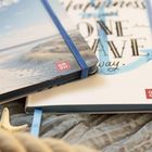 Notizbücher mit den Texten „Happiness is just ONE WAVE away.“ auf einem und „GROH“ Logo auf beiden. Sand und Seestern.