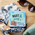 "WORT & weg - Das verrückte Wörterspiel für unterwegs." Eine bunte Metalldose auf Holz mit Sonnenbrille und Schal.