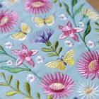 Bestickte Stoffoberfläche mit bunten Blumen und Schmetterlingen auf hellblauem Hintergrund.