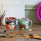 Zwei bunte Tassen: Die linke mit Frida-Kahlo-Porträt, die rechte mit "Viva La Vida", Vögeln und Reh. Pinsel und Farbpalette.