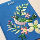 "2021." Ein gestickter Vogel auf blauem Stoff, umgeben von bunten Blumen und Blättern.