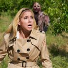 Eine Frau in einem beigen Trenchcoat schaut erschrocken, während hinter ihr eine Zombiegestalt im dichten Grünen auftaucht.