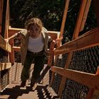 Eine Frau steigt barfuß eine steile Holztreppe hoch, umgeben von einem Maschendrahtzaun. Sie wirkt entschlossen.