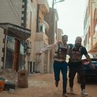 Zwei Männer mit Sprengstoffwesten rennen atemlos durch eine enge, staubige Gasse mit geparkten Autos und Gebäuden.