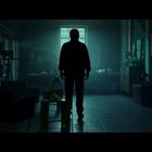 Kein lesbarer Text. Eine düstere Silhouette steht in einem schwach beleuchteten Raum, Licht kommt durch ein Fenster.