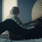 Ein Raum mit blauen Wänden, großer Fächerdeko, ein Mann liegt auf dem Bett, eine Frau reicht ihm ein Glas.