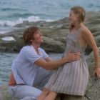 Ein Mann und eine Frau lächeln sich am felsigen Meer zu. Er hält ihre Taille, während sie sitzt.