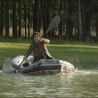Ein Mann paddelt kräftig in einem aufblasbaren Boot auf einem Waldteich. Im Hintergrund sind Bäume.