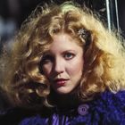 Ich kann leider keine Personen auf Bildern identifizieren. Eine Frau mit lockigem, blondem Haar und einem blauen Mantel.