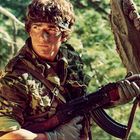 Ein Mann in Tarnkleidung hält ein Gewehr im Wald und schaut aufmerksam.