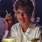 Eine Frau in einem Café hält eine Zigarette und lächelt, Drinks stehen vor ihr.