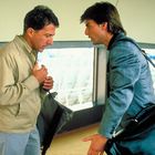 Zwei Männer stehen sich gegenüber, beide mit Taschen; einer trägt eine beige Jacke, der andere eine graue Jacke.